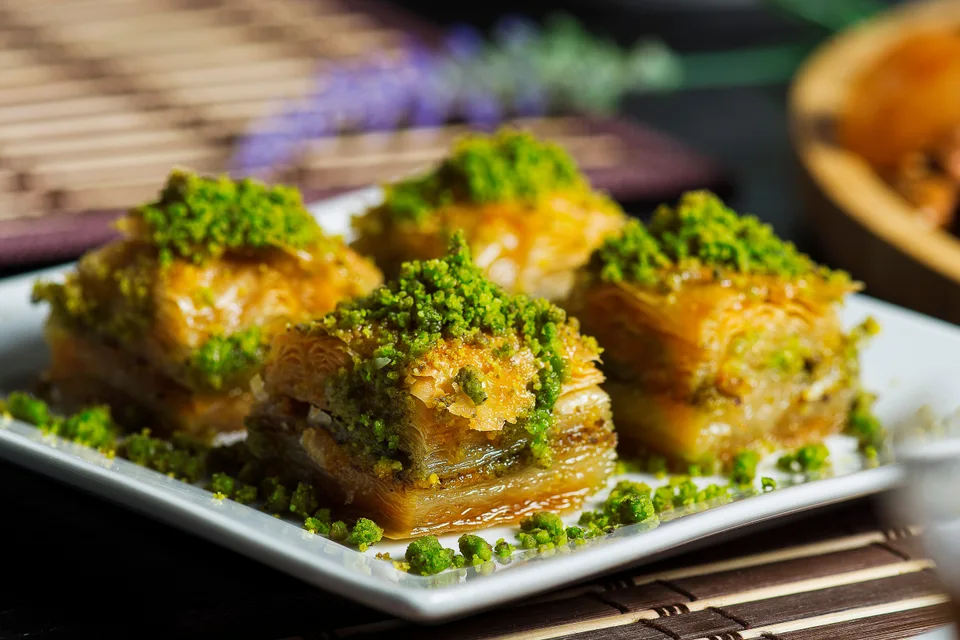 BookNoFees: Ukázková fotografie: Čtyři kousky vrstvené baklavy posypané drcenými pistáciemi na bílém talíři